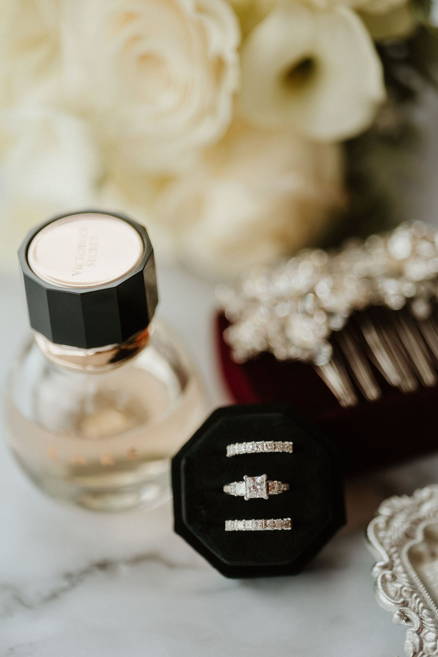 Wedding rings displayed in black box next to perfume bottle on marble surface.