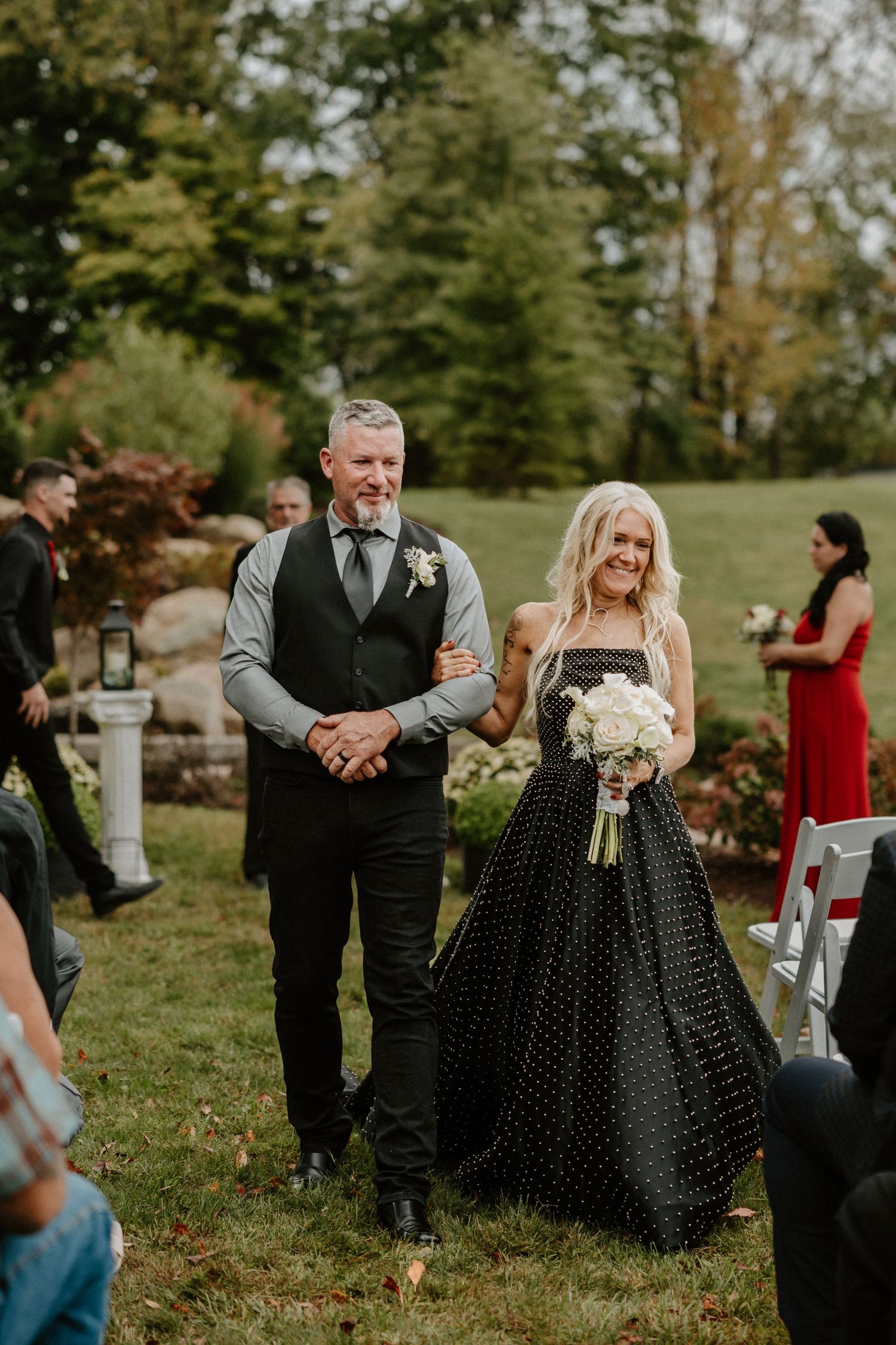 Couple walking down the aisle at outdoor wedding with rock wall and evergreen backdrop.