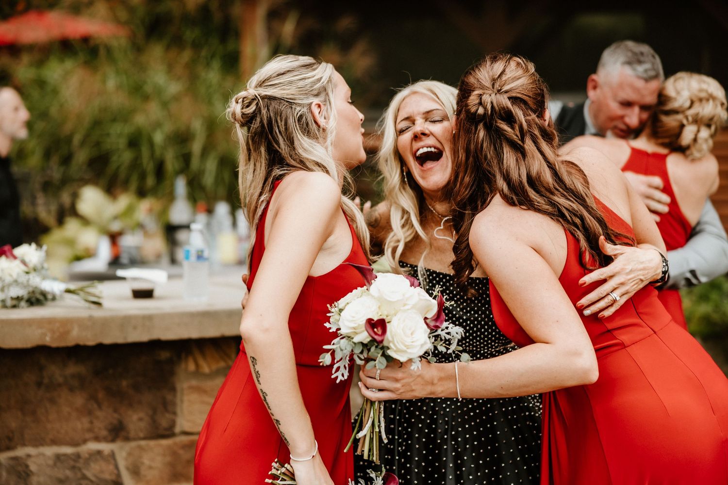 Bridesmaids in red dresses laugh joyfully while celebrating at an outdoor wedding reception.