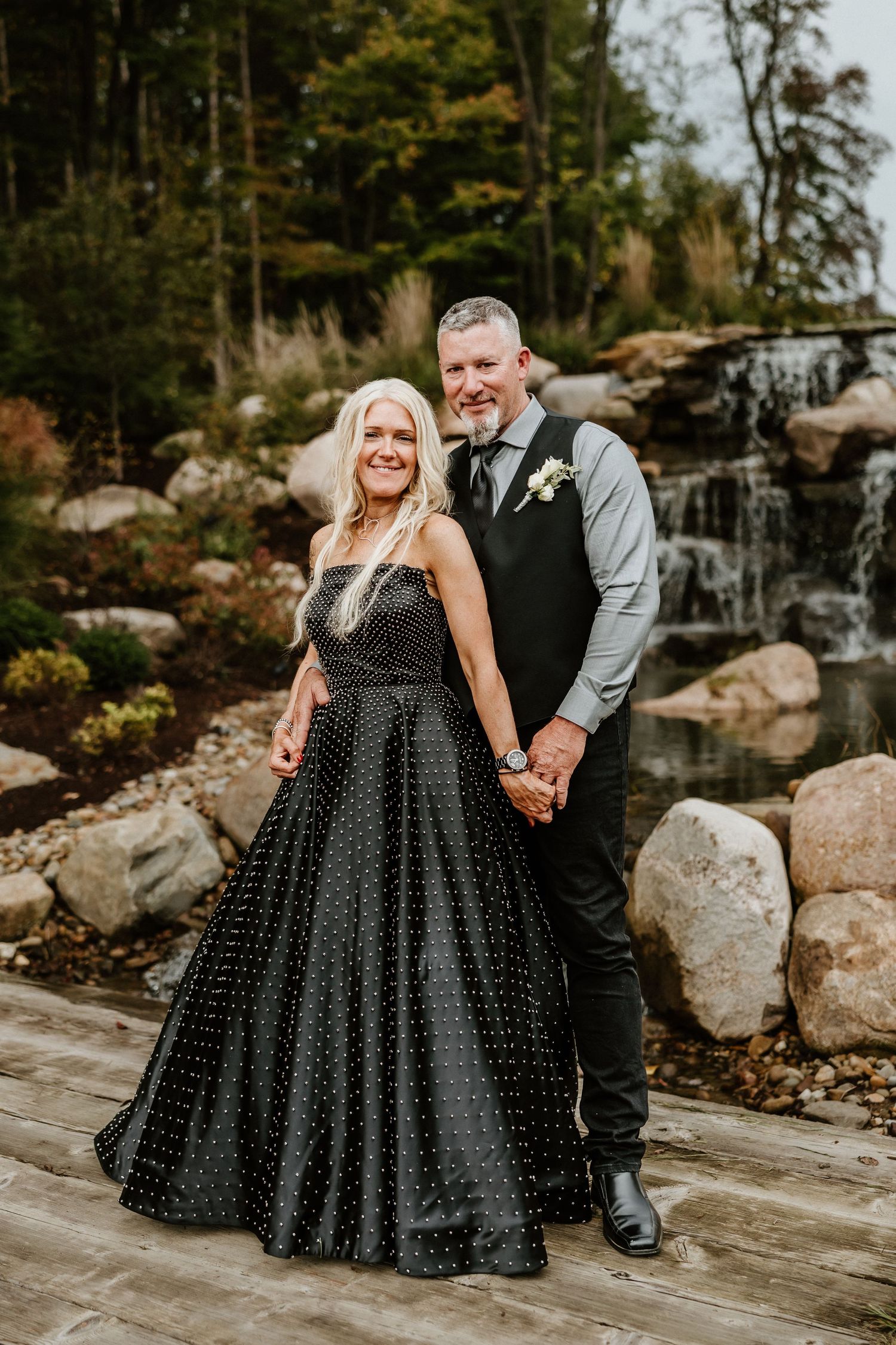 Couple poses by waterfall in formal black dress and grey vest during photo session.