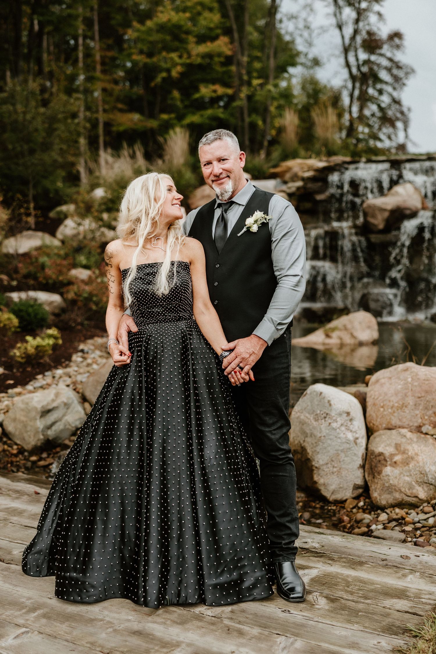 Elegant couple in black gown and grey vest pose against scenic waterfall backdrop.