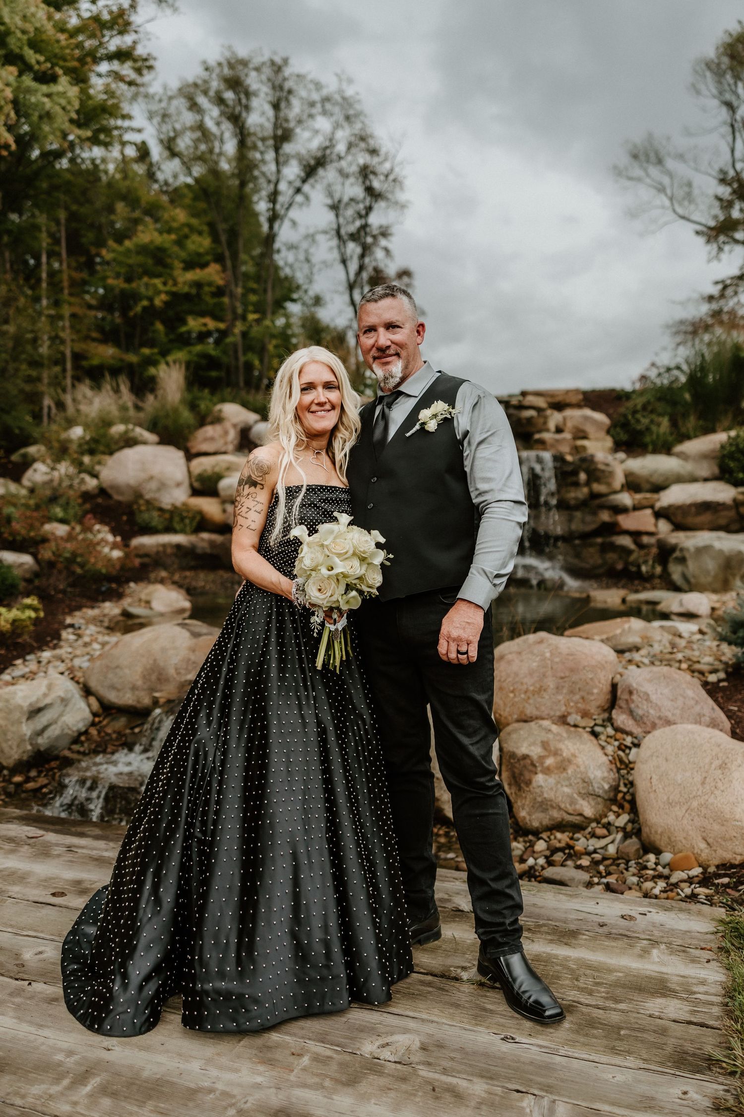 Couple in formal attire holding white bouquet against stone waterfall landscape.