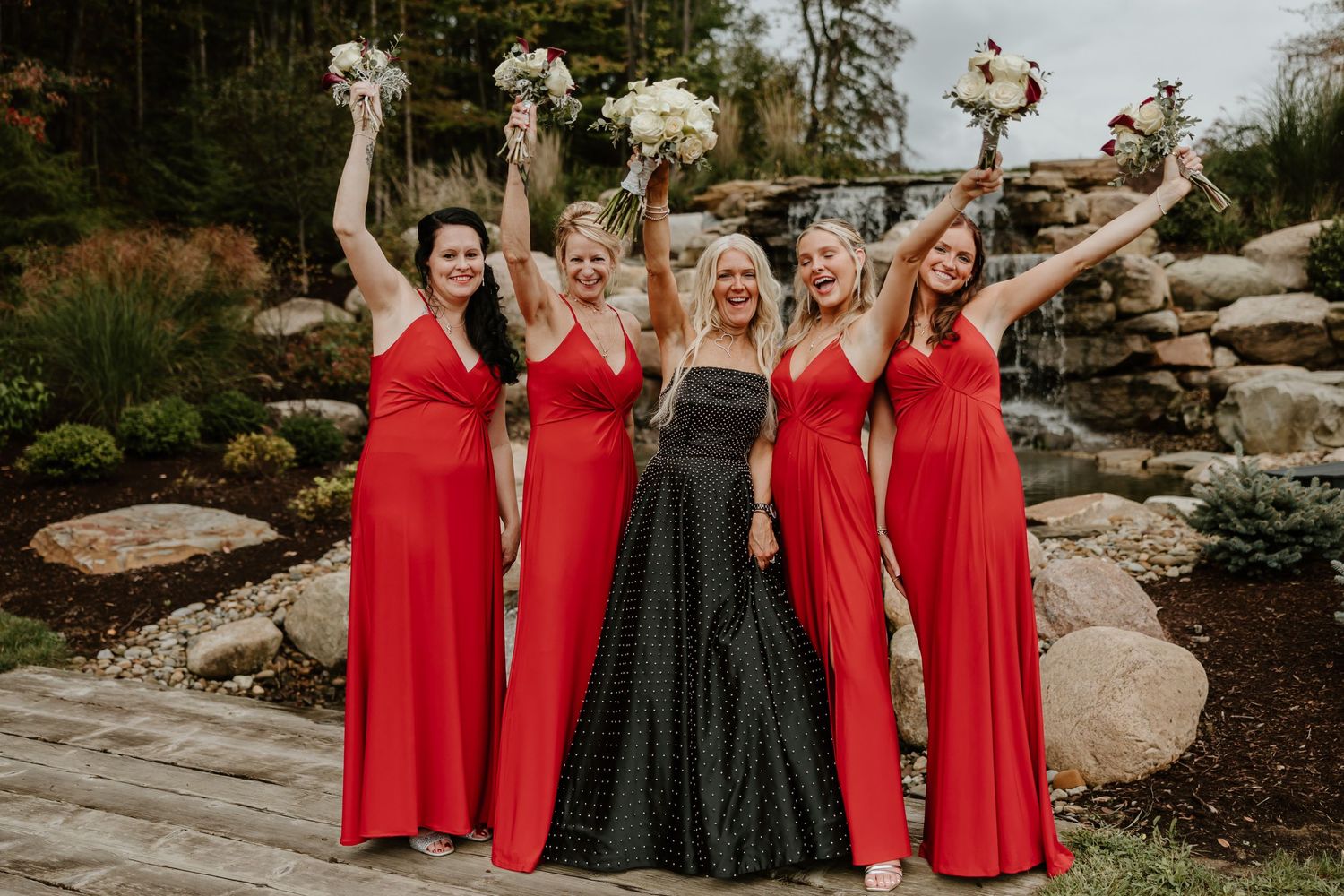 Wedding party in red dresses and black gown celebrate with raised bouquets by waterfall.