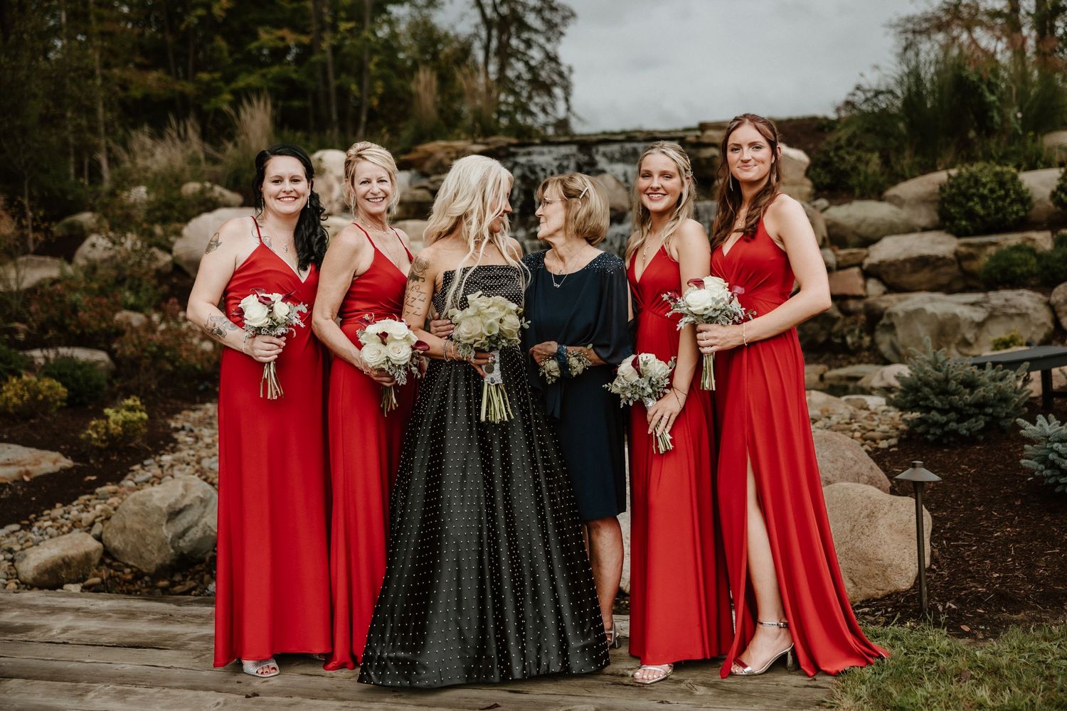 Bridal party in red dresses and black gown gather for photos by rock waterfall.