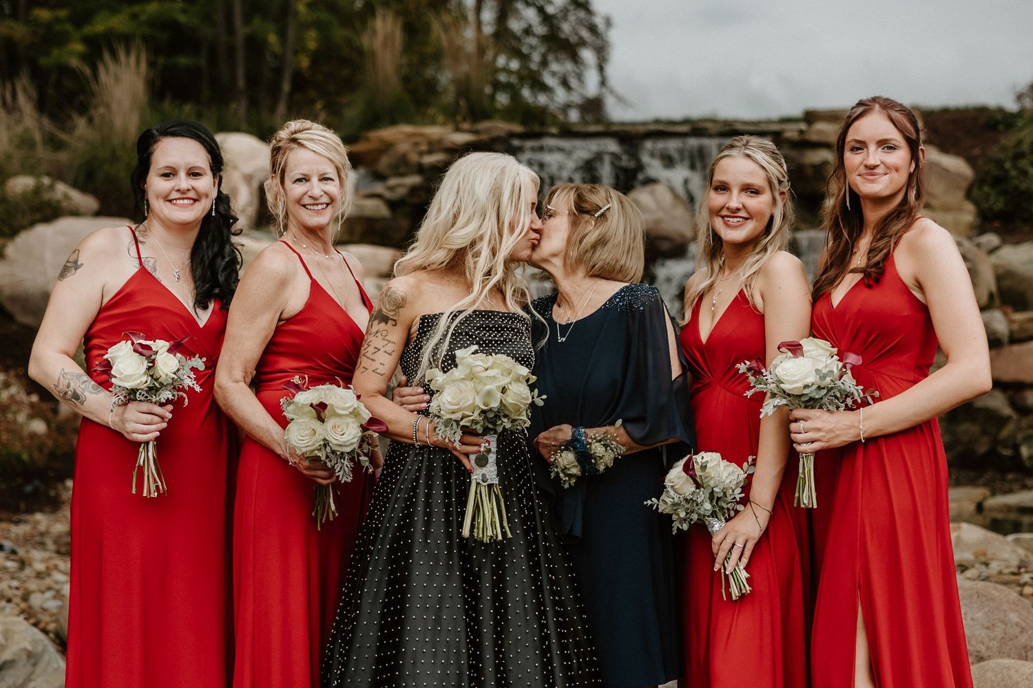 Wedding party with red dresses and black gown share intimate moment by waterfall.