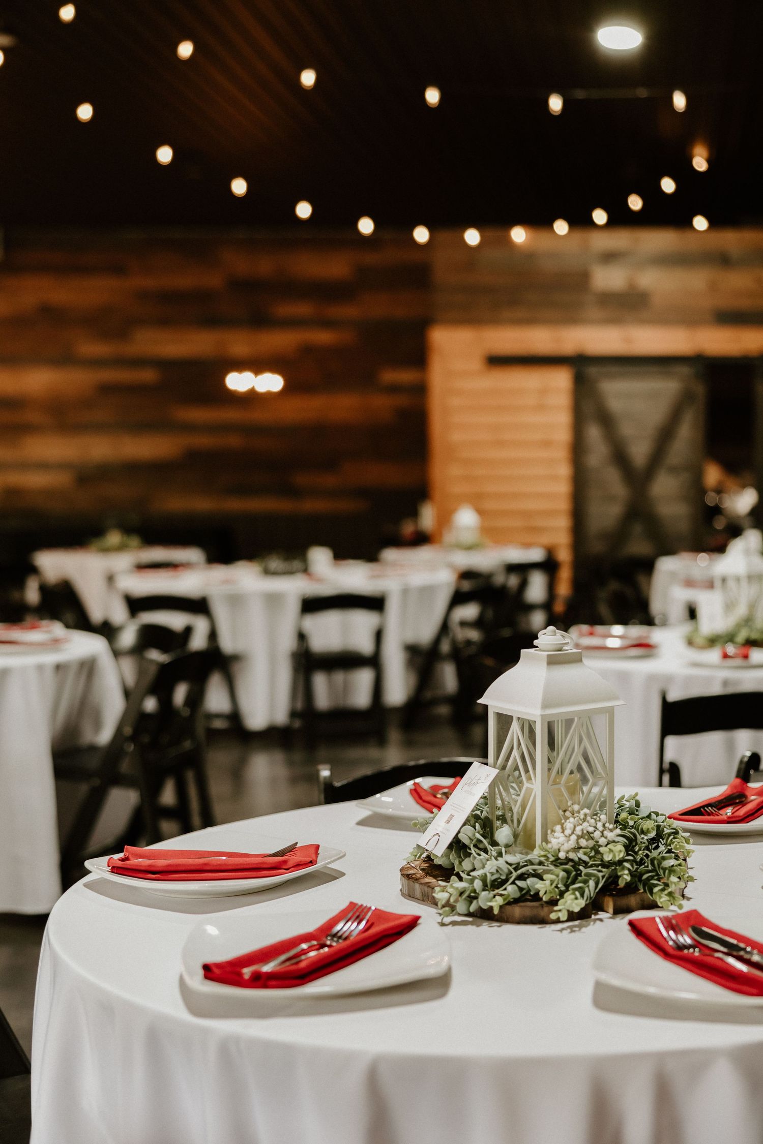 Wedding reception tables set with white linens, red napkins and greenery centerpieces under string lights.