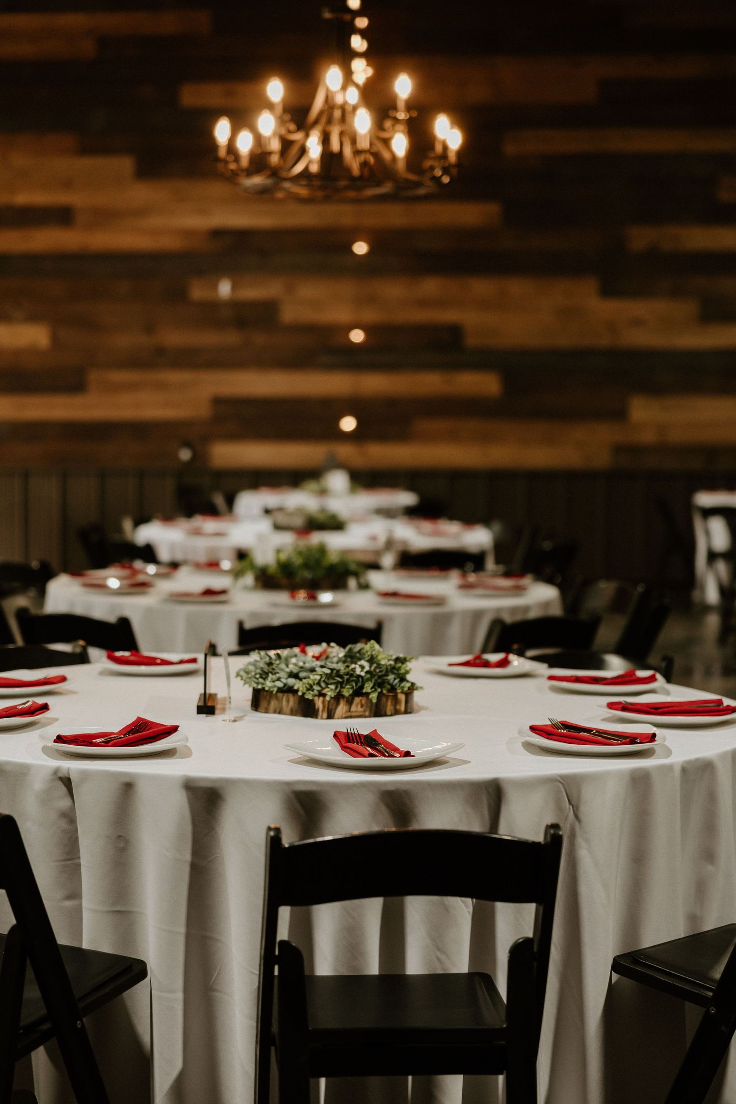 Elegant wedding reception table settings with red napkins and white tablecloths under chandeliers.