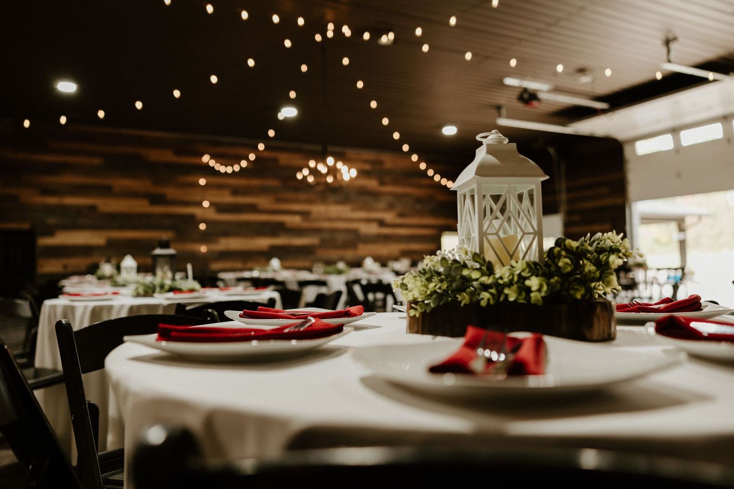 Wedding reception tables with white linens, red accents and lantern centerpieces with string lights overhead.