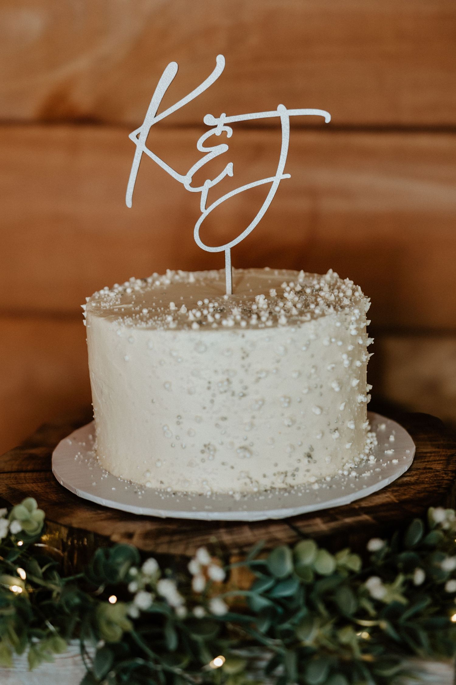 White wedding cake with pearl decorations and monogram topper surrounded by greenery.