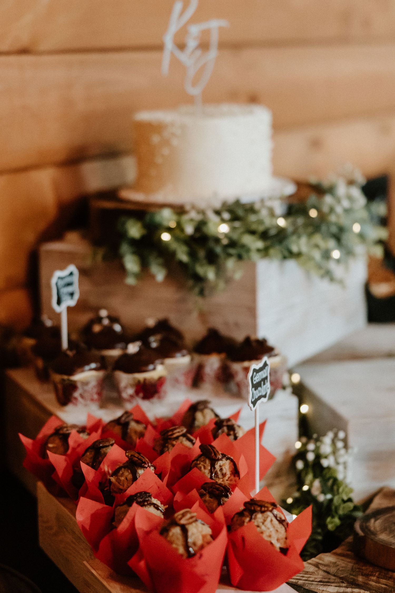Wedding dessert table with white cake and red paper snack containers decorated with greenery.