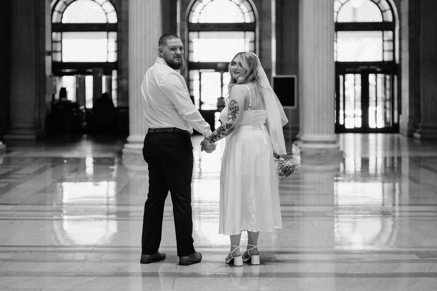 Black and white photo of couple holding hands in grand marble hallway.