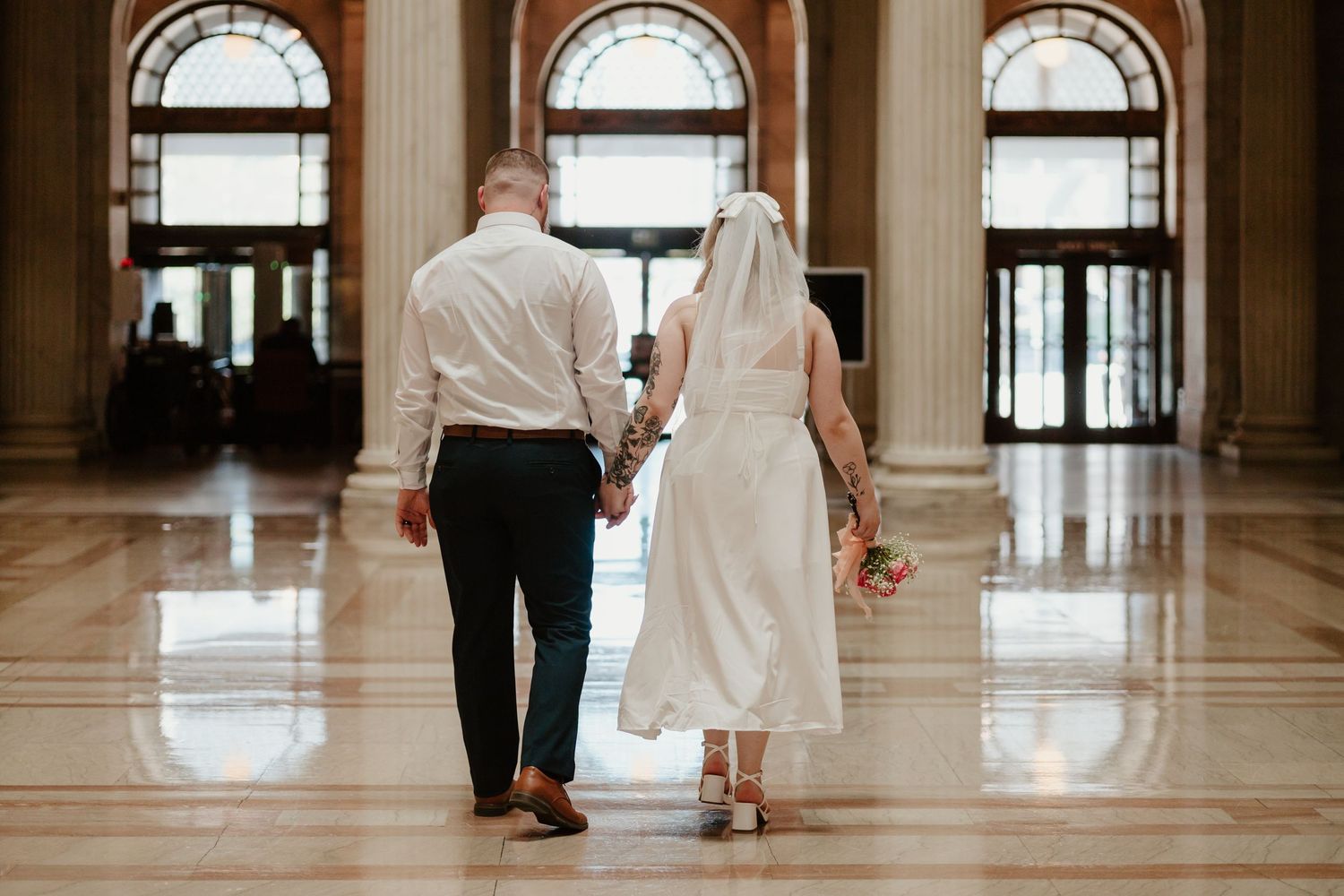 Couple walking away hand in hand down elegant hallway with arched windows.