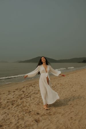 A person in a flowing white dress twirls on a misty beach at dusk with mountains in the background.