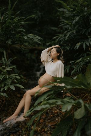Pregnant woman in white crop top sits barefoot on rocks surrounded by lush tropical greenery near a waterfall.