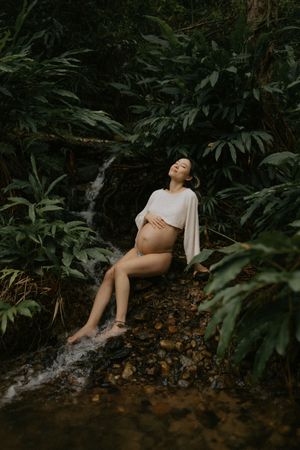 Serene pregnant woman in white wrap top reclines on rocks beside a gentle waterfall in a tropical forest.