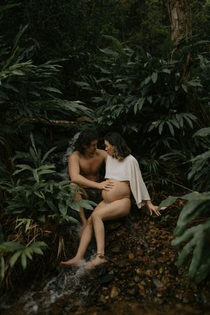 Couple sharing an intimate moment by a jungle stream, woman with baby bump wearing white wrap.