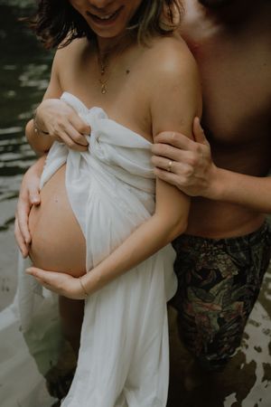 Pregnant woman wrapped in white fabric held by partner during an outdoor maternity photoshoot near water.