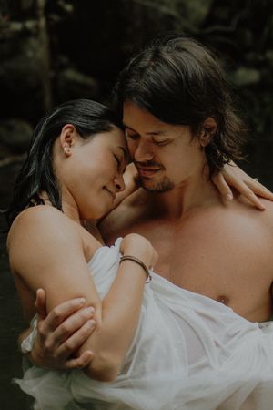 A couple embraces intimately in water, wrapped in white fabric, foreheads touching in a romantic outdoor setting.