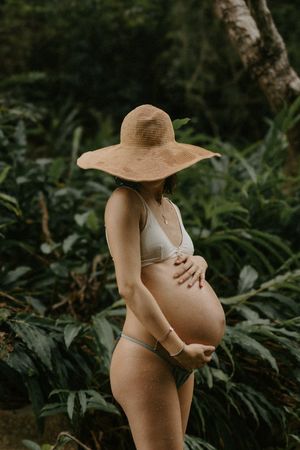 Pregnant woman in white bikini and wide-brim straw hat cradling baby bump in lush tropical garden.