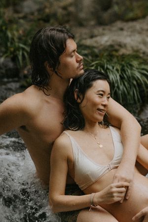 A couple relaxes intimately by a waterfall, woman in white bikini top smiling, man embracing her from behind in nature.