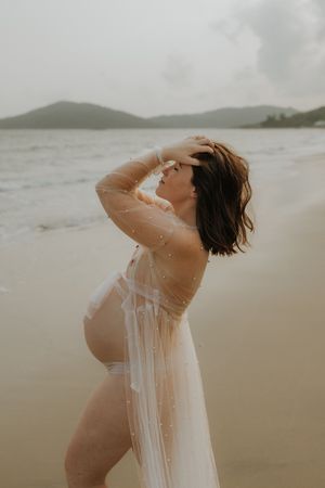 Pregnant woman in sheer white gown poses on beach, hand in hair, with ocean and hills behind her.