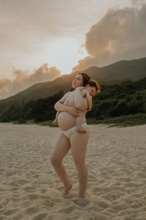 Pregnant woman in white bikini holding child on sandy beach at sunset with green mountains backdrop.
