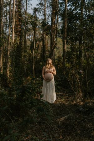 A person in a white dress stands among tall trees in a moody forest setting at dusk.