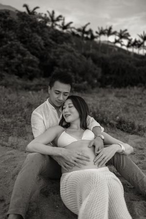 Romantic black and white maternity photo of couple embracing on grassy hillside with palm trees in background.