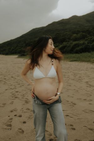 A person in jeans shows their pregnant belly against a dramatic coastal landscape.