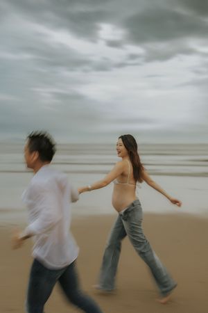 Expecting couple joyfully running and playing together on a moody beach under cloudy skies.