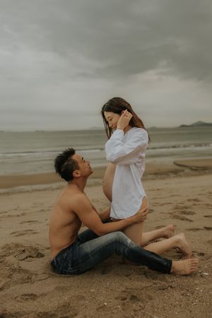 Moody romantic beach photoshoot featuring couple lying in sand with overcast gray sky in background.