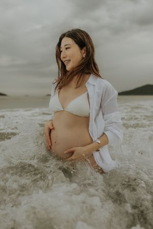 A pregnant woman in a white outfit reclines in ocean waves during a maternity photoshoot at the beach.