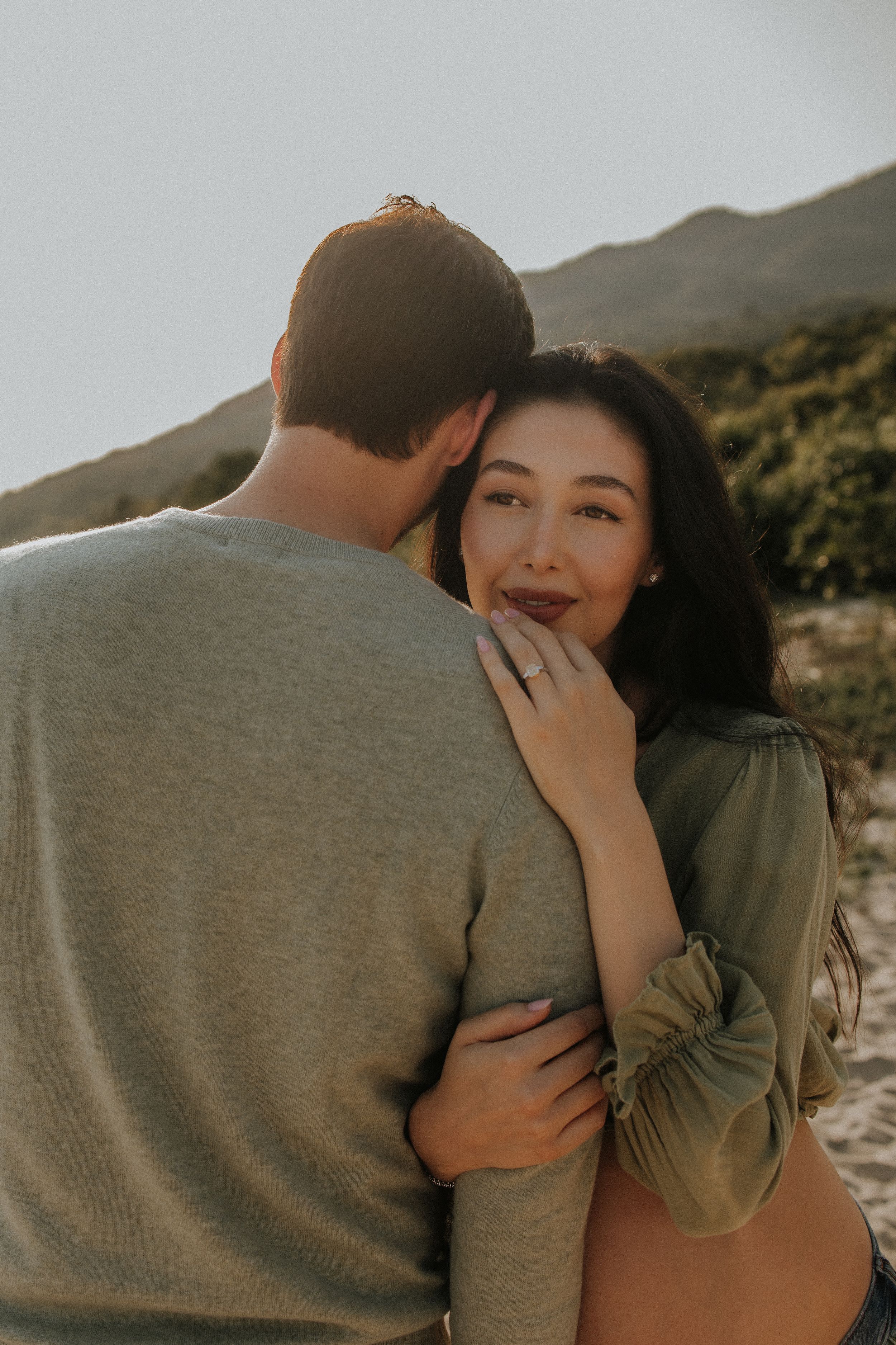 Couple embracing on a beach at sunset, woman in green top smiling over mans shoulder with mountains behind.