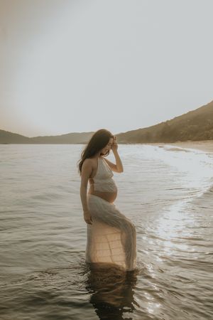 Pregnant woman in white crop top and sheer skirt standing in ocean water at golden hour beach sunset.