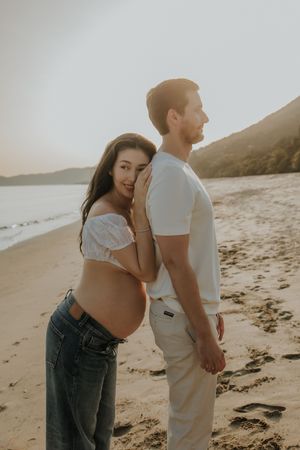 Pregnant woman in white crop top and jeans leans on man's back during golden hour beach maternity photoshoot.