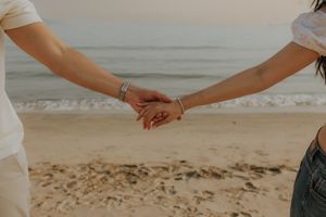 Two people holding hands on a sandy beach at sunset, wearing white tops and silver bracelets.