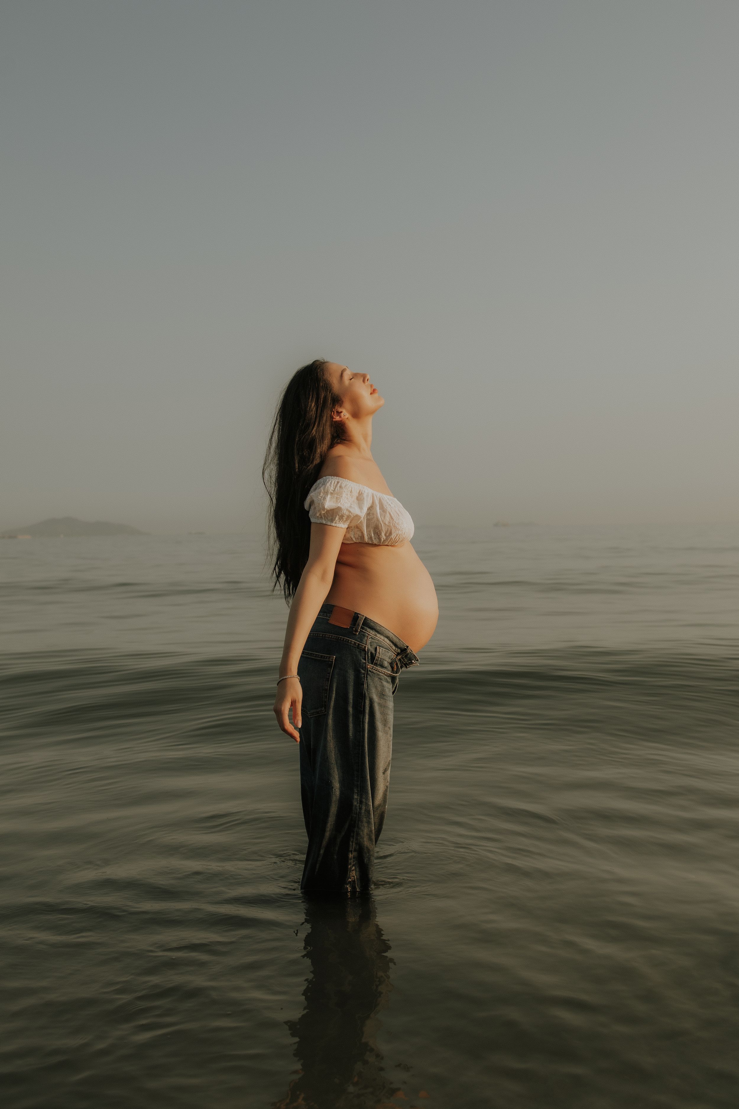 Pregnant woman standing in calm ocean water, wearing white off-shoulder top and jeans, head tilted back at sunset.