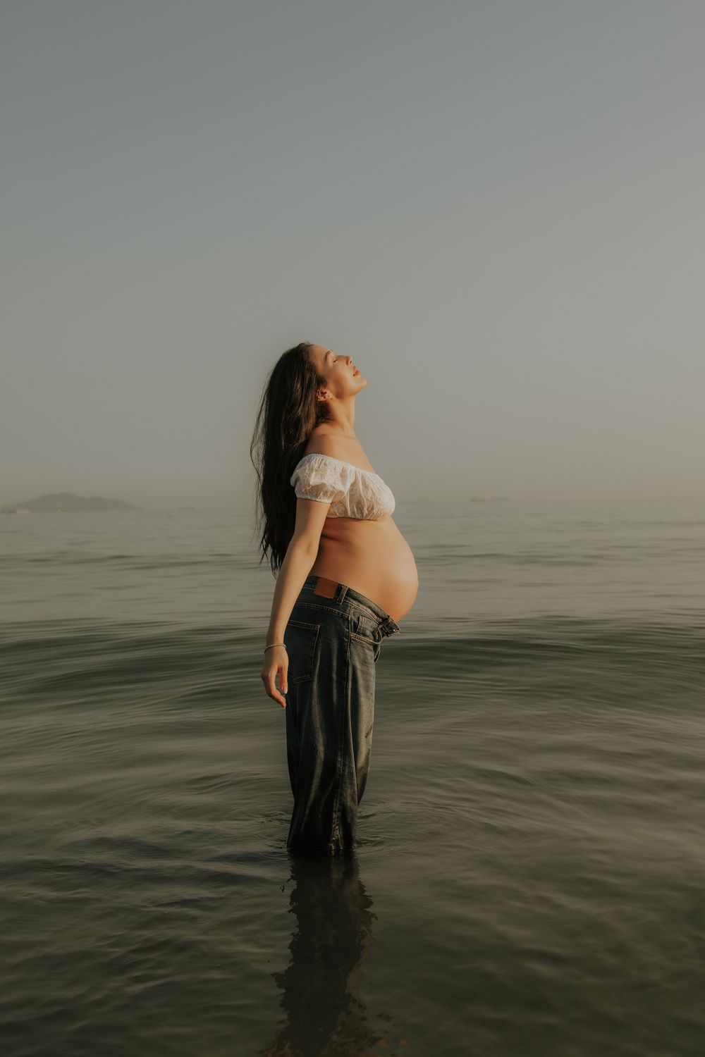 Pregnant woman standing in calm ocean water, wearing white off-shoulder top and jeans, head tilted back at sunset.