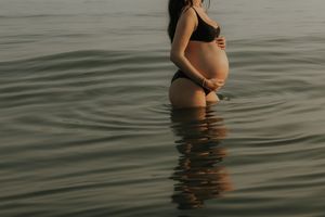Pregnant woman in black bikini standing in calm ocean water, cradling her baby bump at sunset.