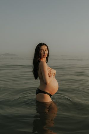 Pregnant woman standing in calm ocean water, cradling her baby bump, with misty mountains in the background.
