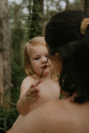 A blonde toddler holds a stick toward a dark-haired woman in a lush green forest setting.