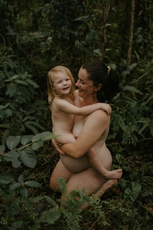 A mother and toddler share a joyful embrace outdoors surrounded by lush green tropical foliage.