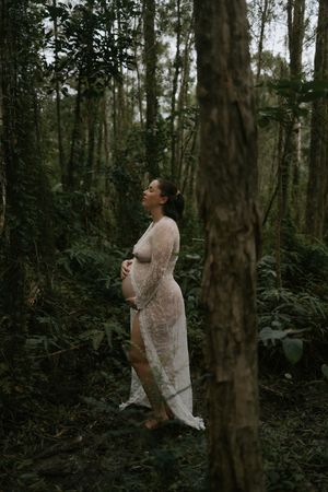 Pregnant woman in sheer white lace gown standing peacefully in lush green forest, cradling her baby bump.