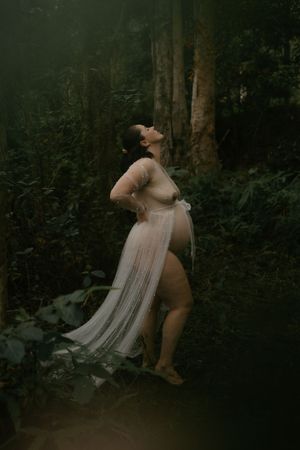 Pregnant woman in sheer white gown poses gracefully in lush green forest setting.
