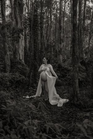 Black and white maternity photo of pregnant woman in flowing white gown standing in lush forest surrounded by ferns and trees.