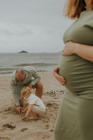 Pregnant woman in green dress holds belly on beach while man and toddler play in sand by the ocean.