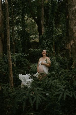 Pregnant woman in white dress smiling serenely among lush green forest foliage and trees.
