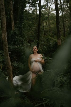 Pregnant woman in flowing white fabric standing in lush green forest during maternity photoshoot.