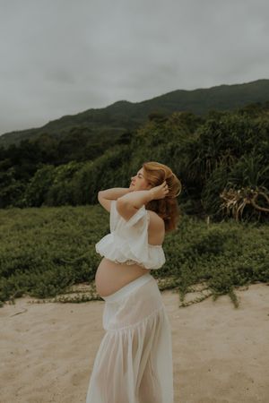 Pregnant woman in white crop top and skirt poses on sandy beach with lush green hills in background.