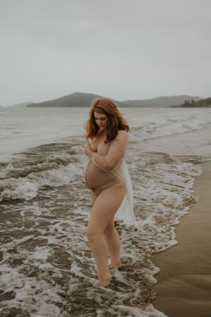 Pregnant woman in sheer wrap standing in ocean waves on a sandy beach with misty hills in the background.